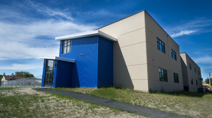 Beige precast concrete and blue metal sided-building with side double door entrance and windows throughout.