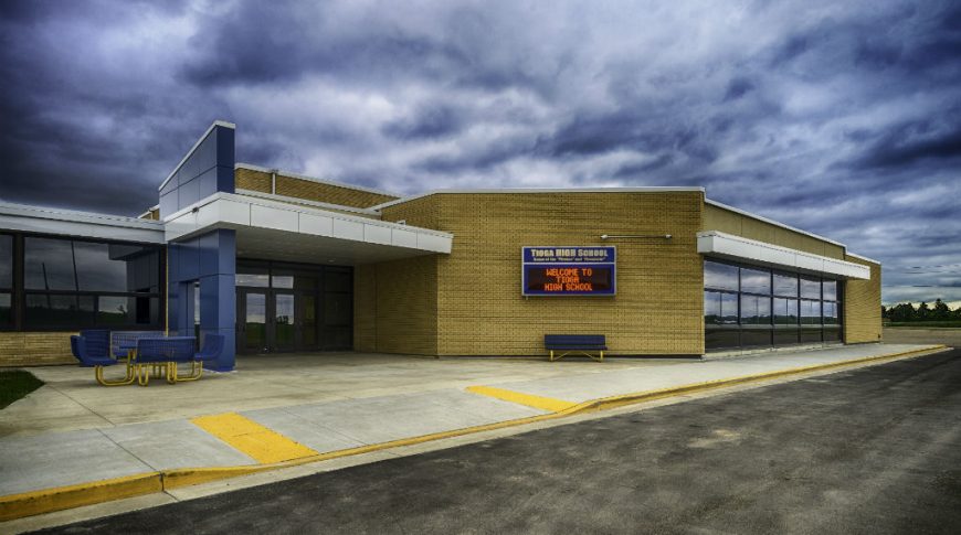 Exterior of yellow bricked high school with blue and silver entryway arches, blue and yellow metal picnic table and bench and digital sign board on the outside of the building.