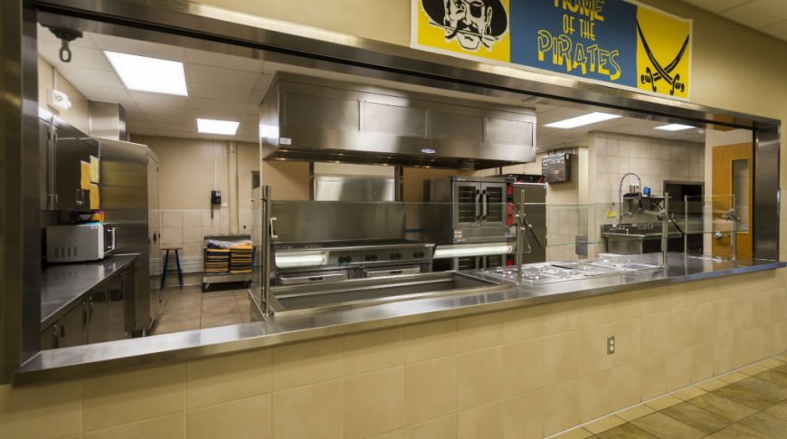 Cafeteria passthrough wall with stainless steel surround and stainless steel kitchen equipment and appliances in the back.