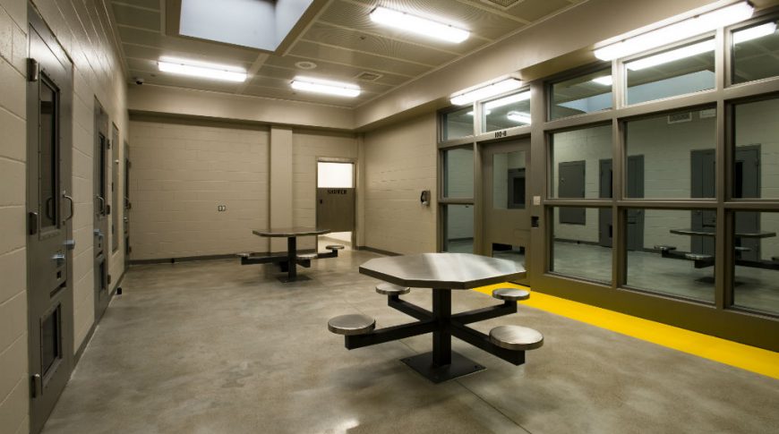 Beige-colored room with concrete floors and two stainless steel and metal tables with surveillance windows to the right and locked doors to the left