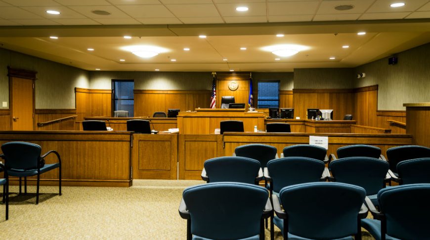 Courtroom with blue chairs for viewers, wooden cabinetry throughout the front with judge's podium and flags and windows on either side