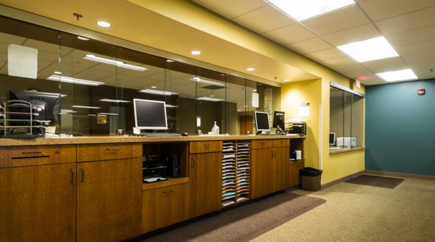 Behind-the-glass view of courthouse support desks with brown cabinetry, speckled brown countertops, brown carpeting and yellow and blue accent walls