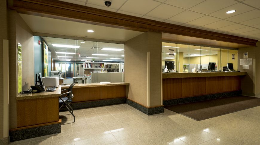 Front reception desks with brown paneling, speckled brown countertops and counter-to-ceiling glass barrier with glimpses of private office areas behind the glass.