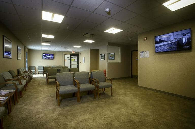 Beige-speckled wall waiting room with beige carpet tiles and chairs throughout with a couple of big-screen TVs on the far right and back walls