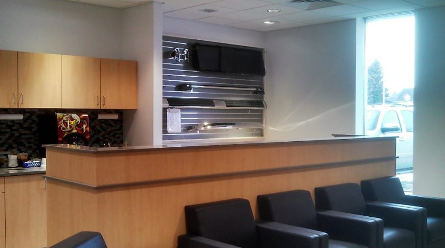 Waiting area with dark gray chairs in the foreground followed by light maple kitchenette cabinetry behind and a sales wall of vehicle audio equipment