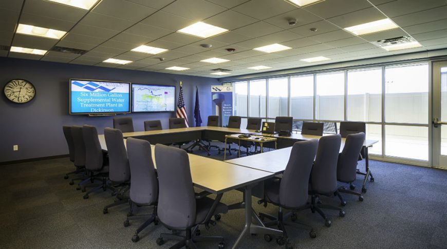 Conference room with dark gray floor squares, square open table to the center of the room with dark gray office swivel chairs around, two large tv monitors to the back wall and wall of windows on the right wall