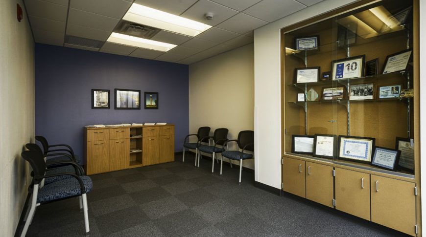Waiting area with dark gray carpeted floor tiles with wood paneled and glass showcase cabinet built in to beige wall to the left and dark blue accent wall to the back with chairs throughout