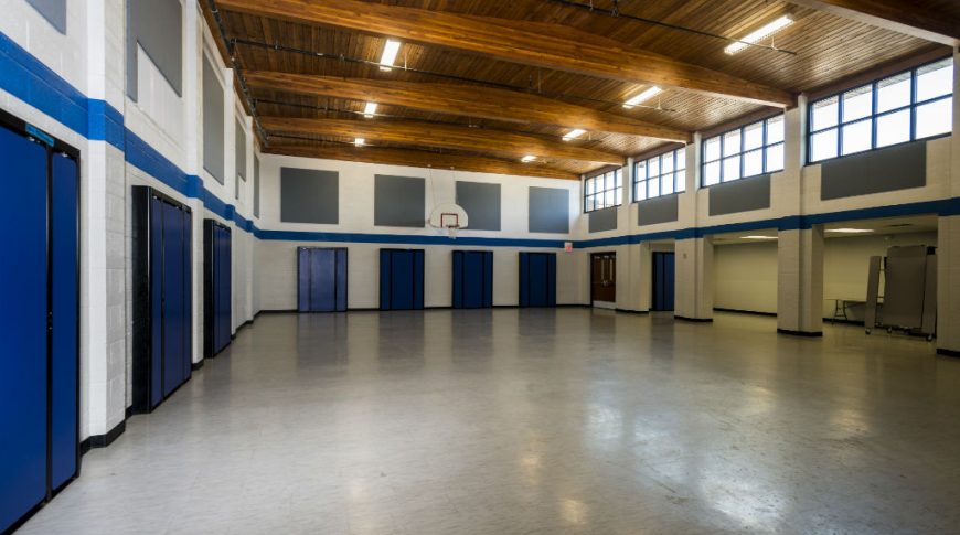 Multi-purpose room with light linoleum tiled flooring with folded rectangular lunch tables along the back and left walls, basketball hoop to the back wall and windows along the upper right wall with wooden-paneled ceiling and exposed trusses