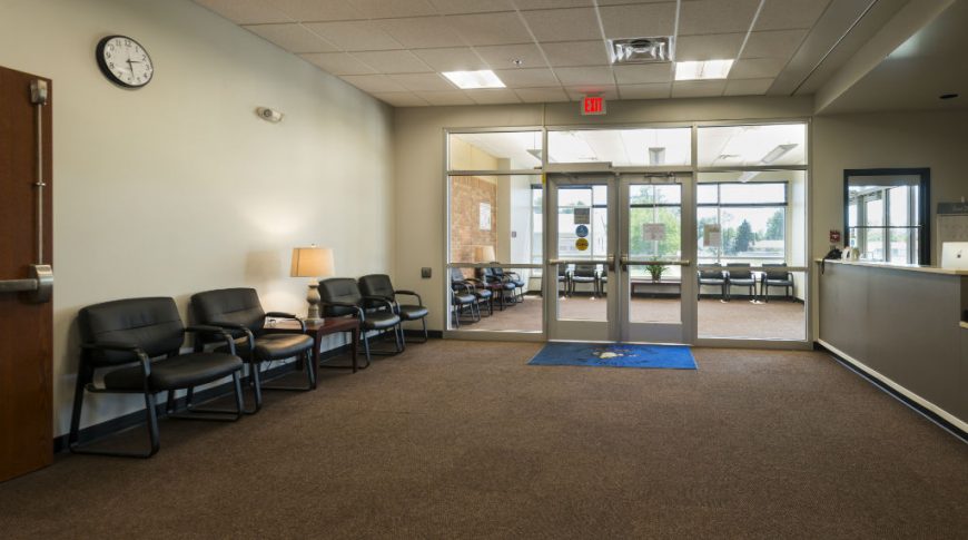 Entrance to school administration offices with large bright windows to the back wall, beige wall to the left with black waiting chairs, and desk station to the right