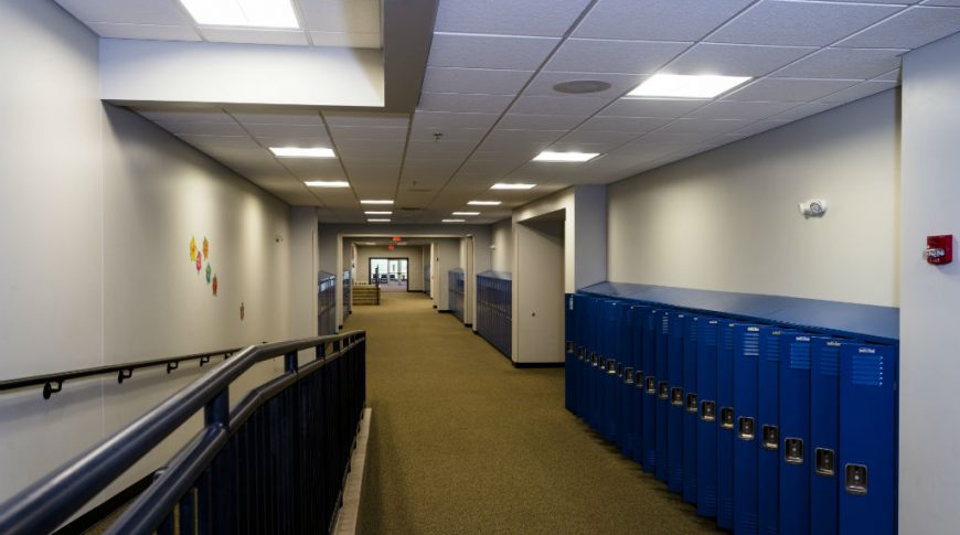 Long hallway of bright blue lockers with light gray walls and ceiling tiles above with brown speckled carpet beneath. and a wheelchair ramp to the left side.