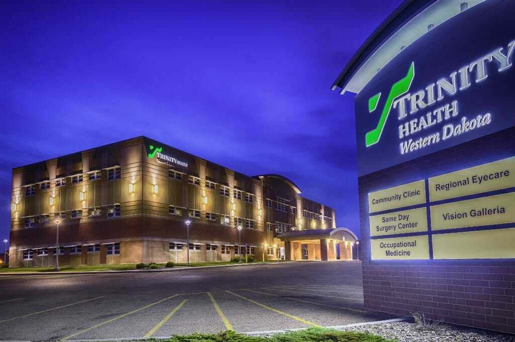 Exterior view of Trinity Health Center in Williston, ND showcasing lit up brown brick façade at dusk with lit up sign to the right in the foreground