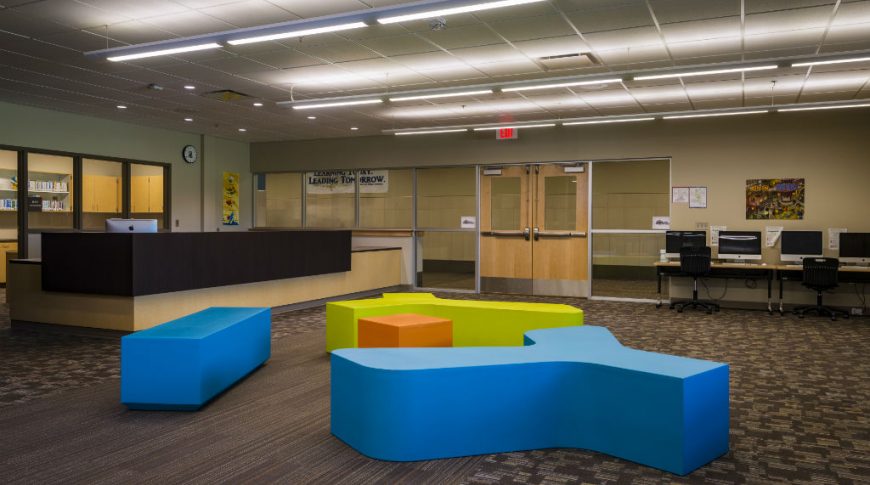 Large room with dark brown floor tiles and wall of windows and doors to the back of the room, with a long maple wood and black countertop reception area to the back left of the room, computer desk stations to the back right of the room and brightly colored blue, lime green and orange geometric bench seats in the middle of the room