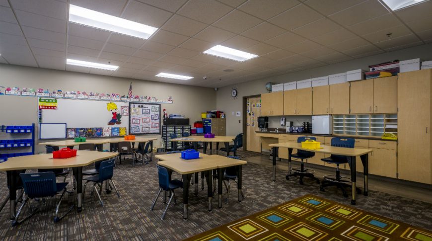 Beige-colored wall classroom with dark brown lined carpet tiles on the floor and ceiling panels with fluorescent lights overhead. Desks scattered about the middle of the room with dark blue plastic chairs, a dry-erase board to the back and light maple cabinetry to the back right of the room
