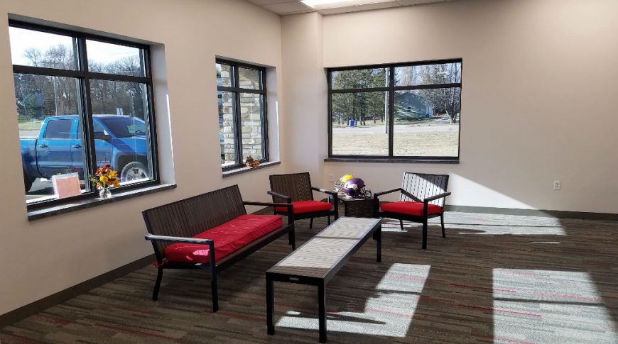Reception waiting area with dark grey and red carpet squares with black and red outdoor furniture couch, tables, and chair, and three windows behind.