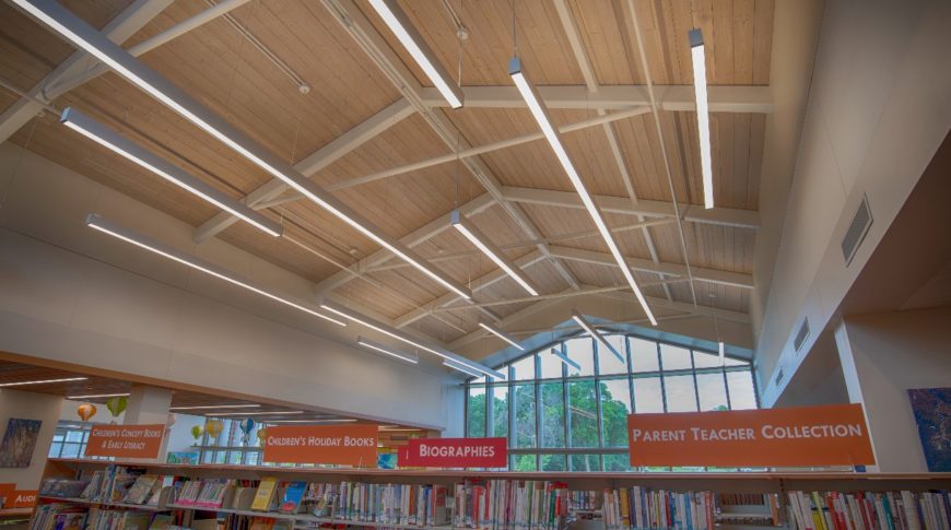 Exposed wood-trussed ceiling in library full of bookshelves, flanked by beige-colored walls and a wall of windows to the rear.