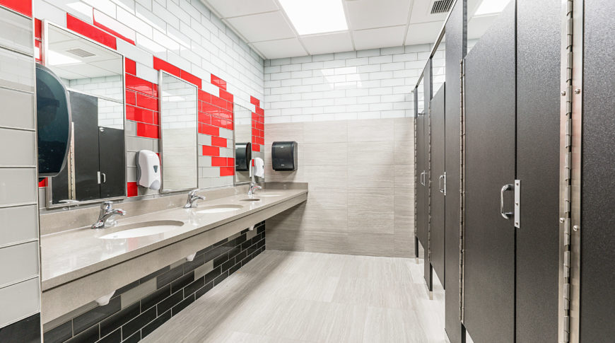Public bathroom with black granite-textured stall doors offset by a wall of subway tiles in gray, red, and black school colors with sinks and mirrors across.