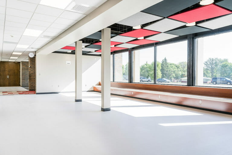 Open commons area with big, bright windows letting sun shine in, a wooden bench across the window sills and school colors in red, black, and grey paneling on the ceiling