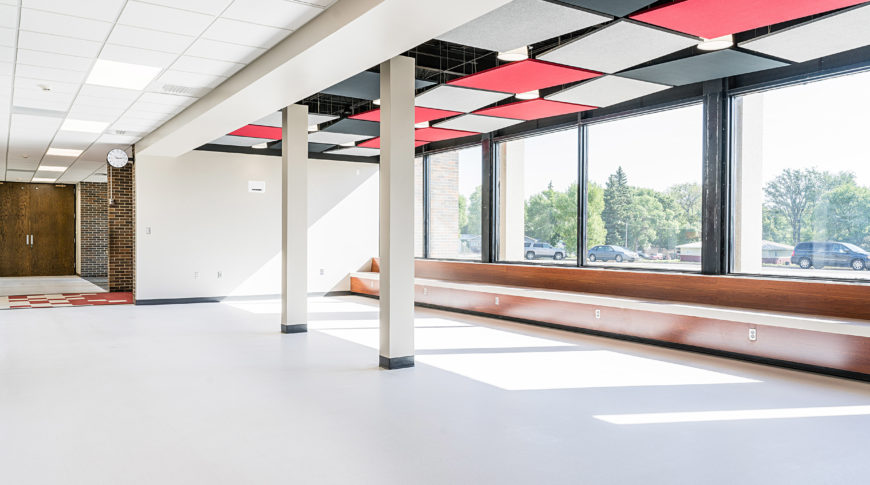 Open commons area with big, bright windows letting sun shine in, a wooden bench across the window sills and school colors in red, black, and grey paneling on the ceiling
