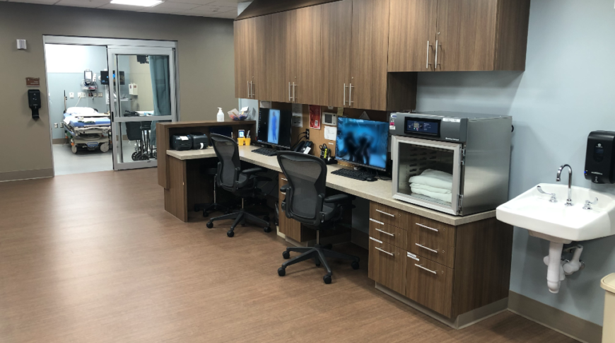 Nurses station with two workstations with computers and chairs and dark wood cabinets, positioned in hallway outside of emergency rooms.
