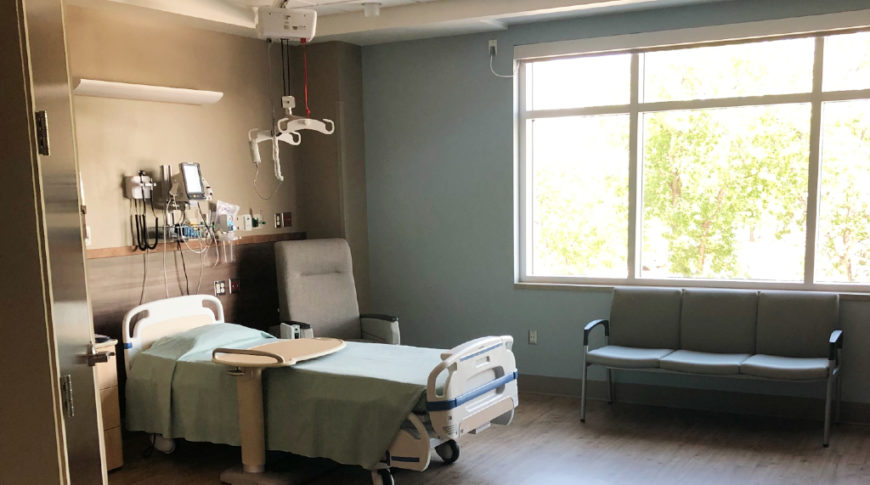 Newly made patient room with robin's egg blue wall on to the back flanked by a tri-panel window and a neutral light brown wall with patient bed and gadgets to the back left of the picture