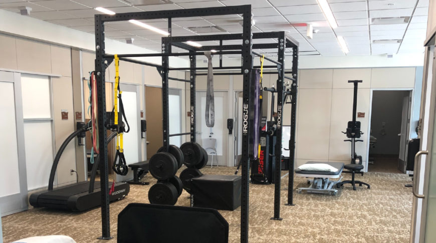 Newly finished therapy and gym room with specialized weight and exercise equipment. Room has neutral brown color palette throughout carpet and walls with bright light from unseen windows.