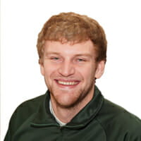 Headshot of a young white man with strawberry blonde hair and scruffy beard smiling with a green polo and white backdrop