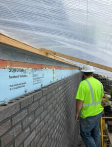 Man in yellow safety shirt standing by partially finished brick outer wall with tarp overhead.