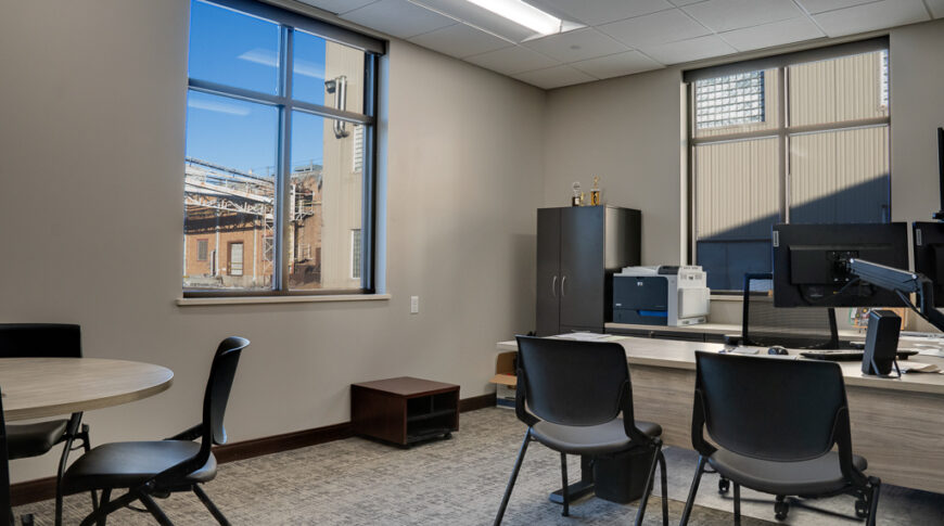 Beige corner office with two large windows on either corner wall overlooking industrial sugar beet plant. U-shaped desk with monitors and printer to the far back right corner and a round table and chairs in the upper left corner
