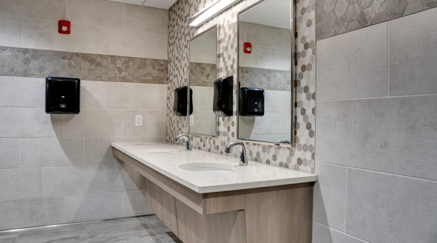 Side view of light weathered stained wood vanity with light quartz top. Two long rectangular mirrors on the back wall with hexagon tiles in gray, beige, and white behind.