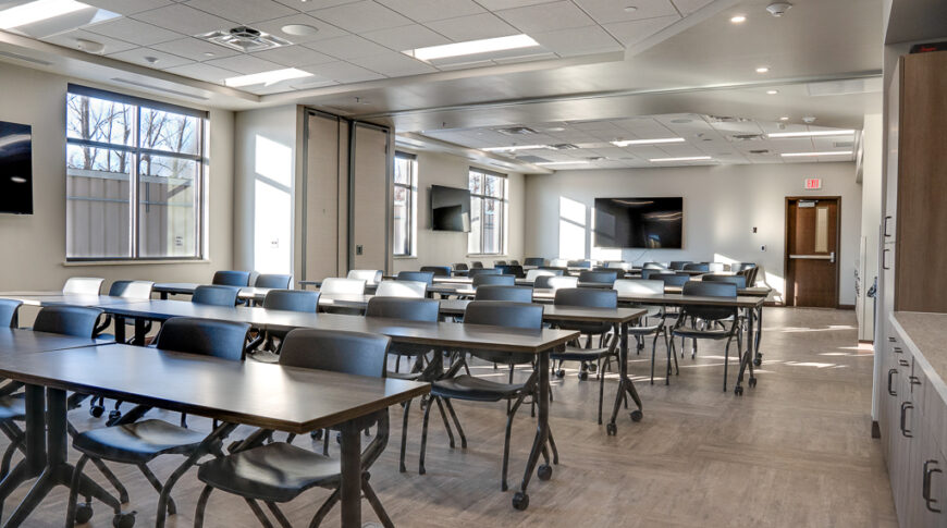 Long conference room with cabinetry on right side, long classroom-style seating down the entire room. Monitors on each wall and large windows on the back left wall.