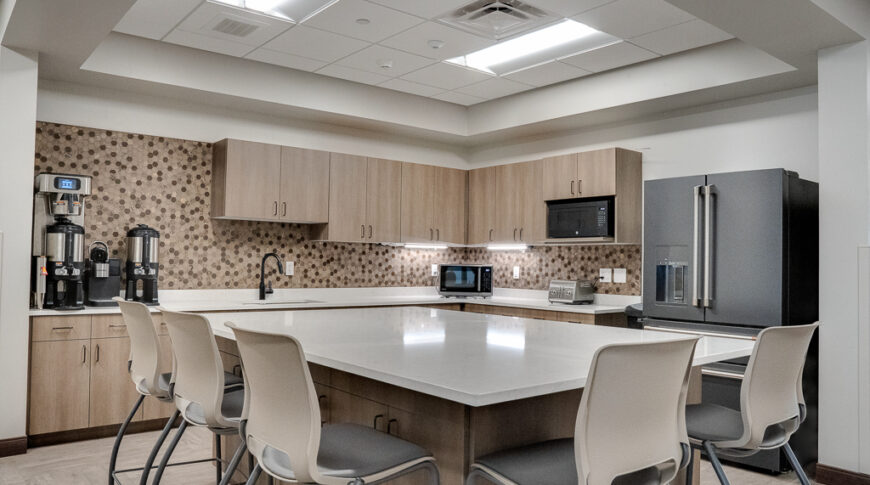 Employee break room kitchen with light weathered wood paneling, stainless steel appliances, and shades of beige hexagon backsplash. Large rectangular island with chair surrounding in the foreground.