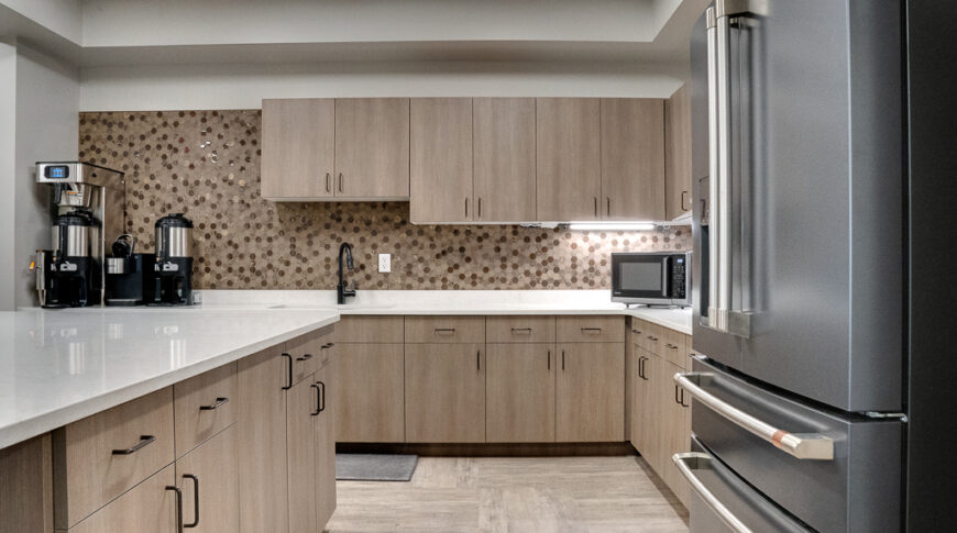 Employee break room kitchen with light weathered wood paneling, stainless steel appliances, and shades of beige hexagon backsplash.