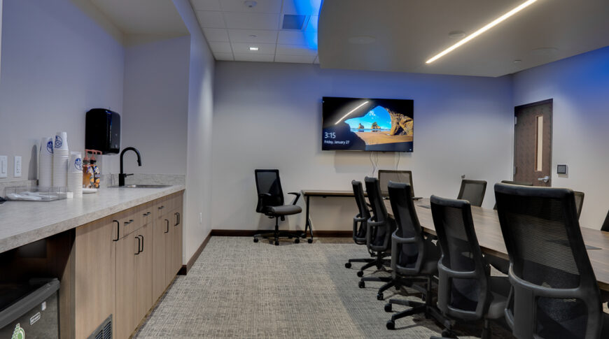 Conference room with long rectangular wooden table in the center surrounded by black office chairs, tv monitor on the back wall, uplight blue panel on the ceiling, and wetbar with cabinetry and minifridge to the left wall.