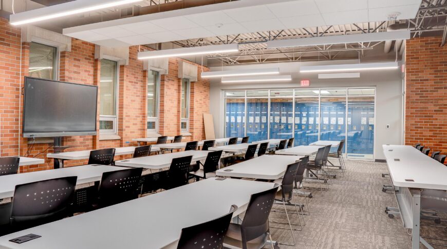Retrofitted classroom space with red brick exterior and interior walls, exposed metal ceiling with modern lights and long round rectangular tables and chairs spanning the length.