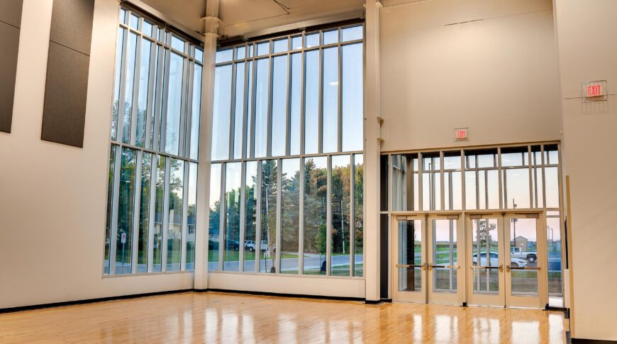 Corner of gymnasium with floor to ceiling two-story windows wrapping around the corner with glass exit doors to the right.