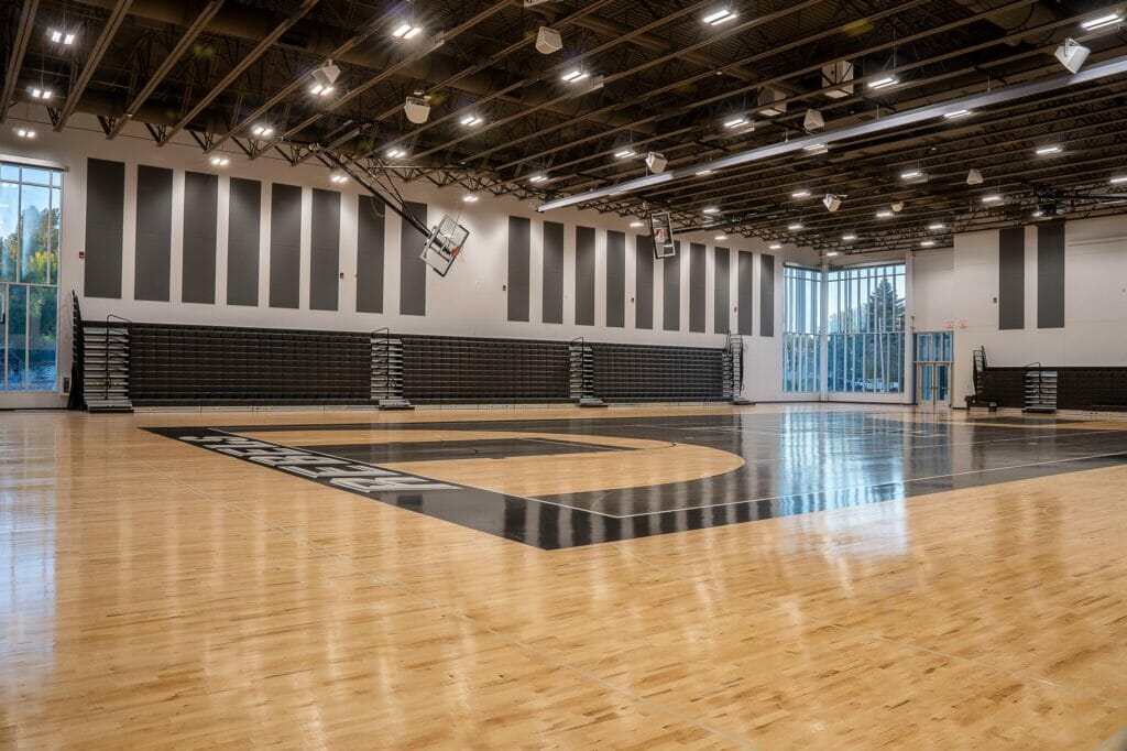 High school gymnasium with bright shiny hardwood floors and black painted basketball court details. Black bleachers folded up on back wall flanked by two-story windows on either side.