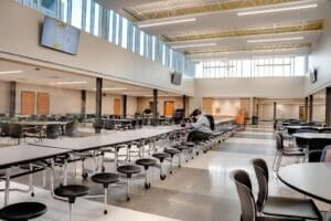 Cafeteria with atrium of windows lining the second story, a row of long rectangular cafeteria tables down the middle with large round tables and chairs on either side.