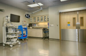 Large medical room with double swinging doors to the back right, cabinetry and sink station to the back left with medical equipment in foreground.