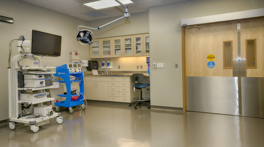 Large medical room with double swinging doors to the back right, cabinetry and sink station to the back left with medical equipment in foreground.