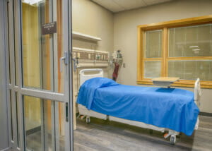 Prep and Recovery patient room with sliding glass doors and yellow privacy curtain with blue-patient bed in foreground and large maple windows on back wall.