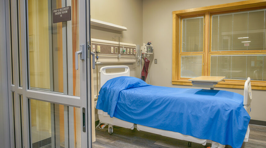 Prep and Recovery patient room with sliding glass doors and yellow privacy curtain with blue-patient bed in foreground and large maple windows on back wall.