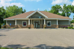 Parking lot exterior view of InVision eye clinic with green shaker siding, white and stone columns and brown shingled roof.