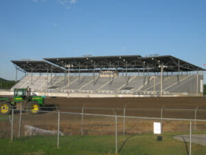 Exterior view of North Dakota State Fair Grandstands from far opposite side of the field on sunny day with blue sky in background and green tractor in foreground.