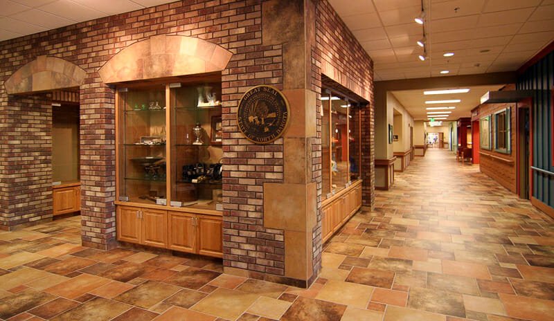 Corridor of two hallways meeting with shades of brown and beige tiles on the floor with faux brick walls with oak cabinetry on each side.