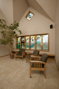 Tiled seating area with three wooden chairs with dark brown cushions, stained glass windows behind and faux birch tree in a pot to the far left.