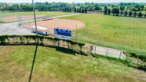 Intersection of two outdoor baseball fields with fence in between them. Lush green grass and trees outlining the fields.