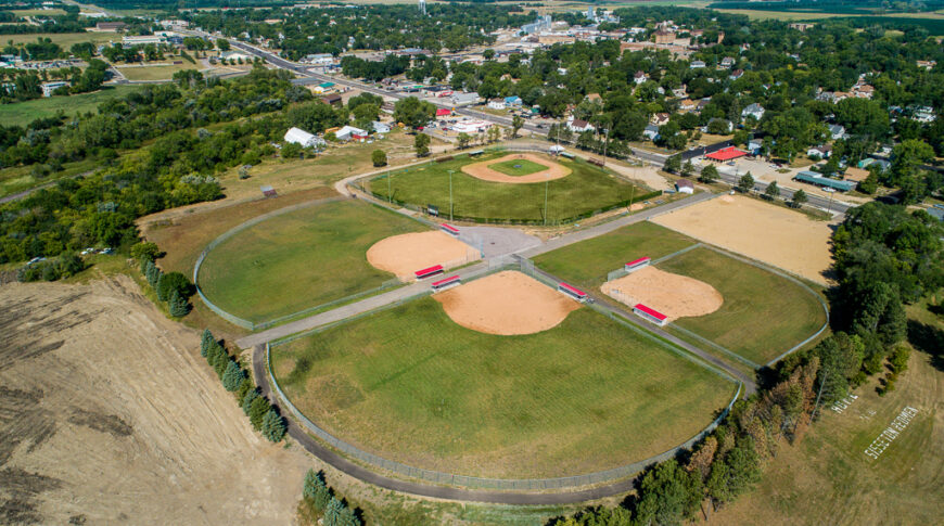 Aerial view of four baseball fields on sunny summer day with lush green trees surrounding and small city in the background.