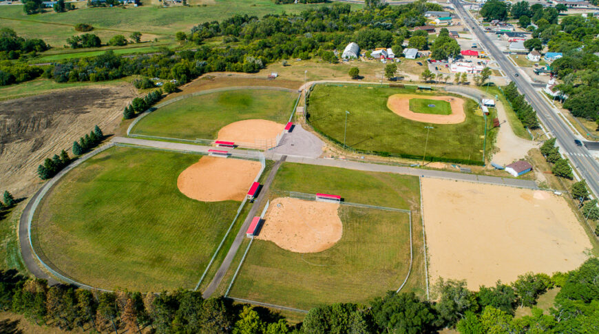 Aerial view of four baseball fields on sunny summer day. Three newer fields have bright red-roofed dugouts and older field has bleacher system.
