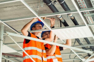 Two men wearing blue hard hats and orange and white reflective safety vests working on piping behind industrial ceiling panels.