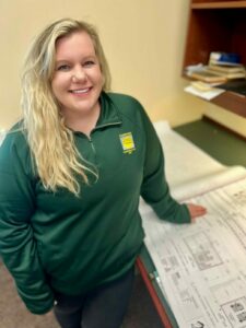 White woman with long bright blonde wavy hair in green long-sleeve Comstock Construction polo leaning next to desk with construction blueprints.